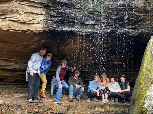 A group of nine people poses under a rocky overhang with a waterfall cascading behind them.