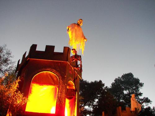 A spooky Halloween scene featuring a castle with a skeleton and a ghostly figure atop it, illuminated by orange light.