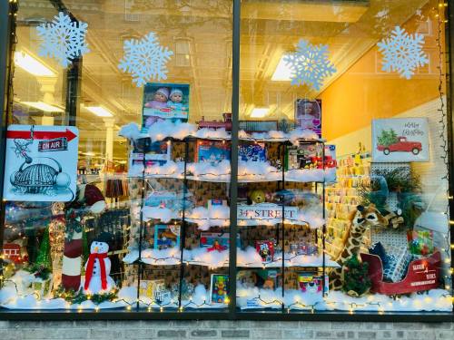 A festive store window display with toys, snow decorations, and holiday lights, featuring a snowman and a giraffe.