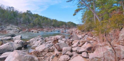 A serene river flows through a rocky landscape, surrounded by lush green trees under a clear blue sky.