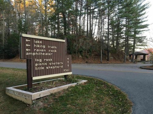 Wooden signpost with directional arrows for lake, hiking trails, amphitheater, and picnic shelters, surrounded by trees.