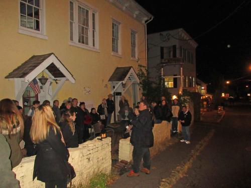 A group of people gathered outside a yellow building at night, listening to a speaker near a street with lights.