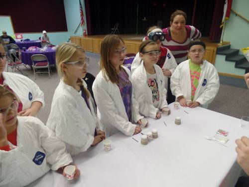 A group of children in lab coats and goggles stands at a table, engaged in a science activity.