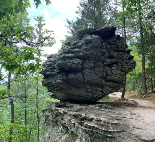 A large, balanced rock sits atop a cliff surrounded by trees and a dirt path in a natural landscape.