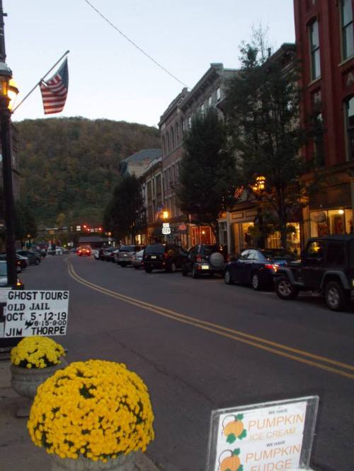 A quaint street lined with shops and cars, featuring autumn decorations and a backdrop of hills at dusk.