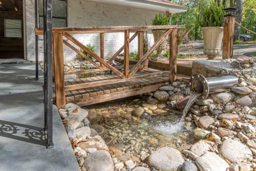 A wooden bridge over a rocky stream, with water flowing from a metal pipe into a clear pool.
