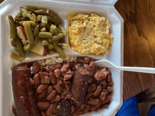 A styrofoam container with rice, red beans, sausage, green beans, and a cheesy potato dish.