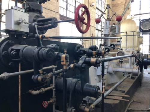 Close-up of vintage industrial machinery with pipes, valves, and gauges in a large, well-lit space.