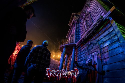 A spooky house illuminated with colorful lights, with people observing in the foreground at CreepyWorld attraction.