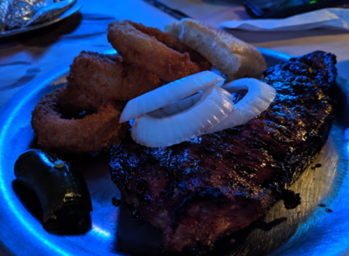 A plate of grilled steak topped with onion slices, accompanied by onion rings and a jalapeño pepper.