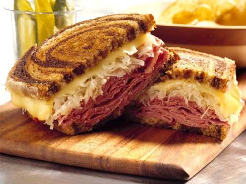 A sliced sandwich on rye bread filled with corned beef, sauerkraut, and melted cheese, served with pickles.