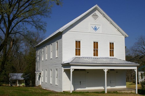 A white, two-story building with a Masonic symbol on the front, surrounded by trees and a clear blue sky.