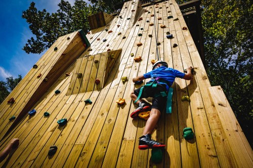 A child climbs a wooden climbing wall, wearing a helmet and harness, surrounded by trees and blue sky.