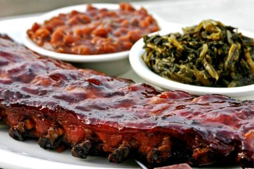 A plate featuring glazed ribs, baked beans, and collard greens in white bowls.