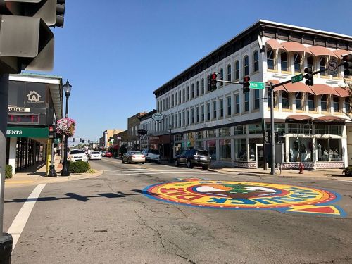 A colorful mural on the street at an intersection, with shops and traffic signals in a small town setting.