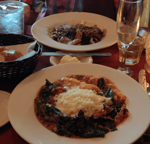 A table setting with two plates of pasta, one with spinach and cheese, and the other with a dark sauce, plus drinks.