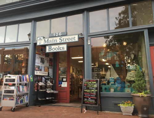 A cozy bookstore entrance with large windows, displaying books and decorative items, on Main Street.