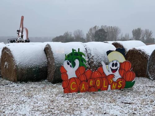 A colorful sign with cartoon ghosts and pumpkins stands near snow-covered hay bales in a winter landscape.