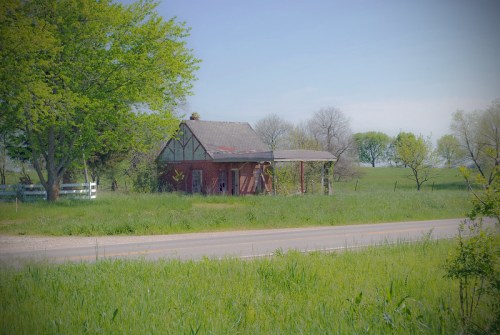 An old, weathered red house surrounded by green grass and trees, near a quiet road on a sunny day.