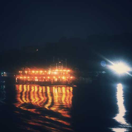 A dimly lit boat on water at night, reflecting warm lights against a dark backdrop of hills.