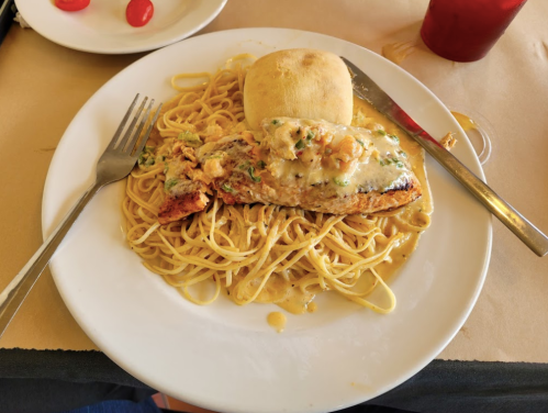 A plate of creamy pasta topped with a baked fish fillet and a small bread roll, with a drink in the background.
