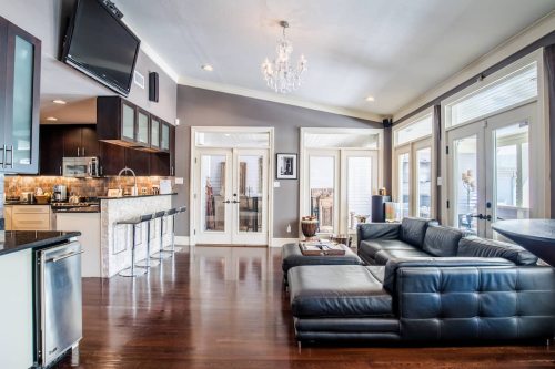 Modern living room with a black leather sectional, a chandelier, and a kitchen bar with stools. Bright, open space.