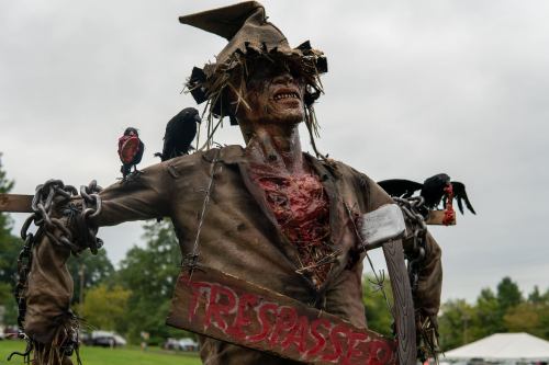 A creepy scarecrow figure with a torn outfit, bloodied appearance, and crows perched on its shoulders, holding a "Trespasser" sign.
