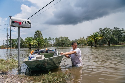 A man stands in floodwaters, pulling a boat near a mailbox marked 7376, with dark clouds and palm trees in the background.