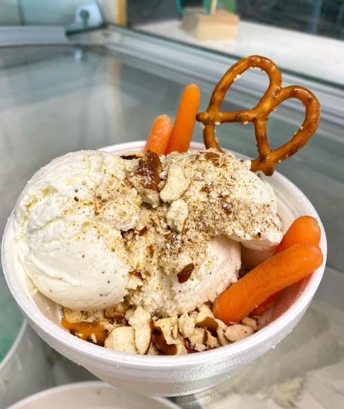 A cup of ice cream topped with crushed nuts, pretzel, and carrot sticks, served in a white container.