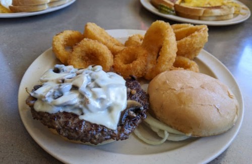 A plate with a cheeseburger topped with mushrooms, a soft bun, and a side of crispy onion rings.