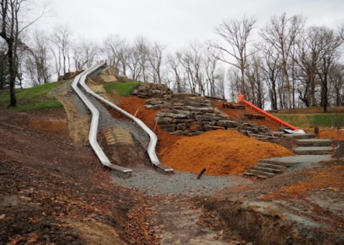 Two slides on a hillside, surrounded by bare trees and gravel paths, with piles of mulch and rocks nearby.