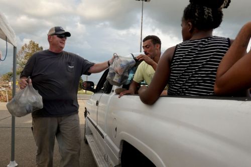 A man hands a bag of food to a person in a truck, while another person sits nearby, under a cloudy sky.