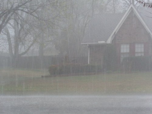 A house is partially obscured by heavy rain, with blurred trees and a wet lawn in the foreground.