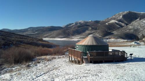 A green yurt on a snowy landscape, surrounded by mountains and a lake under a clear blue sky.