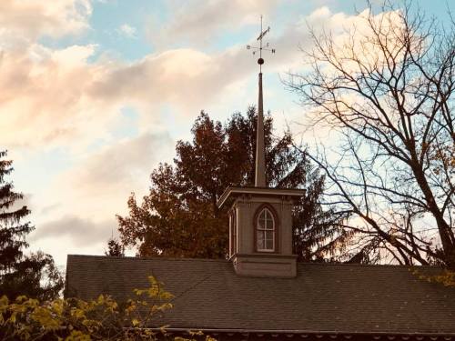 A church steeple with a weather vane against a backdrop of trees and a colorful sky.
