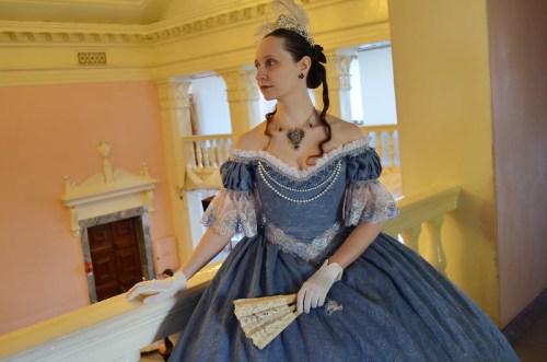 A woman in a vintage blue gown and tiara stands elegantly by a balcony, holding a fan.