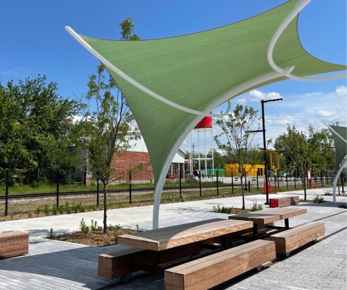 A shaded outdoor seating area with wooden benches and green canopies, surrounded by trees and a clear blue sky.