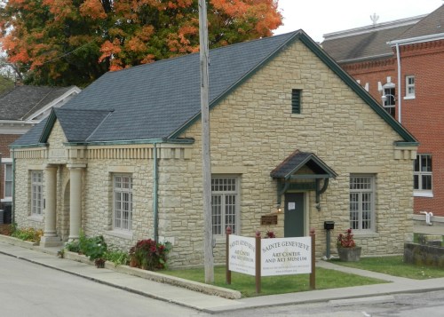A stone building with a green roof, featuring large windows and a sign for an art center and museum. Autumn trees in background.