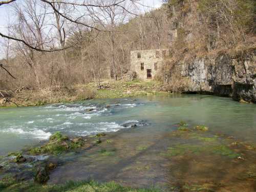 A serene river flows past a stone building, surrounded by trees and rocky cliffs under a clear sky.