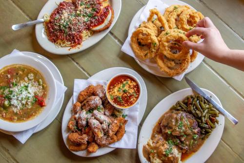 A spread of various dishes including spaghetti, onion rings, fried chicken, green beans, and a bowl of soup on a table.