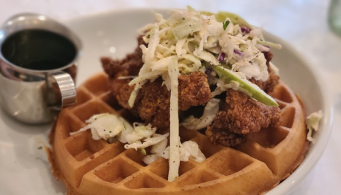 A golden waffle topped with crispy fried chicken and coleslaw, served with a small cup of syrup.