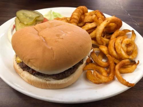 A plate with a hamburger topped with mustard, pickles, and lettuce, accompanied by curly fries and a slice of tomato.