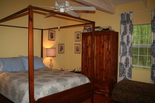 Cozy bedroom with a four-poster bed, lamp, wooden wardrobe, and patterned curtains, featuring warm yellow walls.