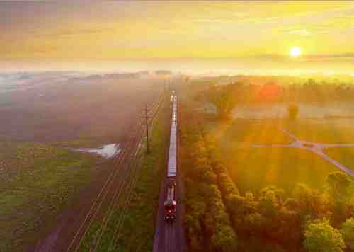 A train travels along a misty landscape at sunrise, with golden light illuminating the fields and trees.