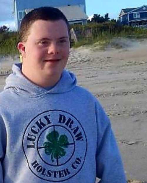 A young man with Down syndrome smiles while standing on a beach, wearing a gray hoodie with a shamrock design.