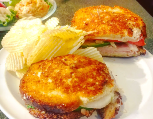 A crispy sandwich with layers of meat and vegetables, served with a side of potato chips on a white plate.