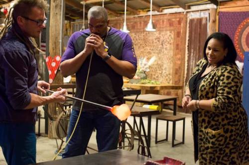 A glassblower demonstrates glassblowing techniques to two intrigued onlookers in a workshop setting.