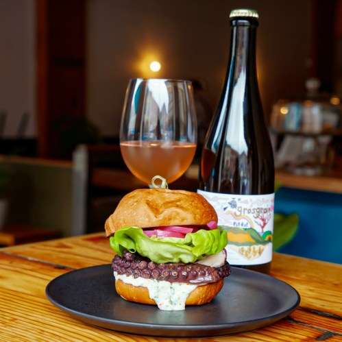 A gourmet burger with octopus, lettuce, and sauce on a black plate, accompanied by a glass of wine and a bottle.