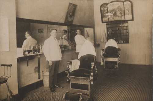 A vintage barbershop interior with two barbers and two clients in barber chairs, featuring a mirror and shelves of products.