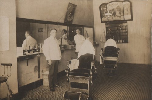 A vintage barbershop interior with two barbers and two clients in barber chairs, featuring a mirror and shelves of products.